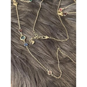 K&ouml;nnte beinhalten: Eine zarte, goldfarbene Halskette mit mehreren Str&auml;ngen. Die Halskette verf&uuml;gt &uuml;ber verschiedene farbige Edelsteine, darunter Blau, Rot und Gr&uuml;n, die entlang der Kette angeordnet sind. Der Verschluss und der Anh&auml;nger sind sichtbar und zeigen das filigrane Design.