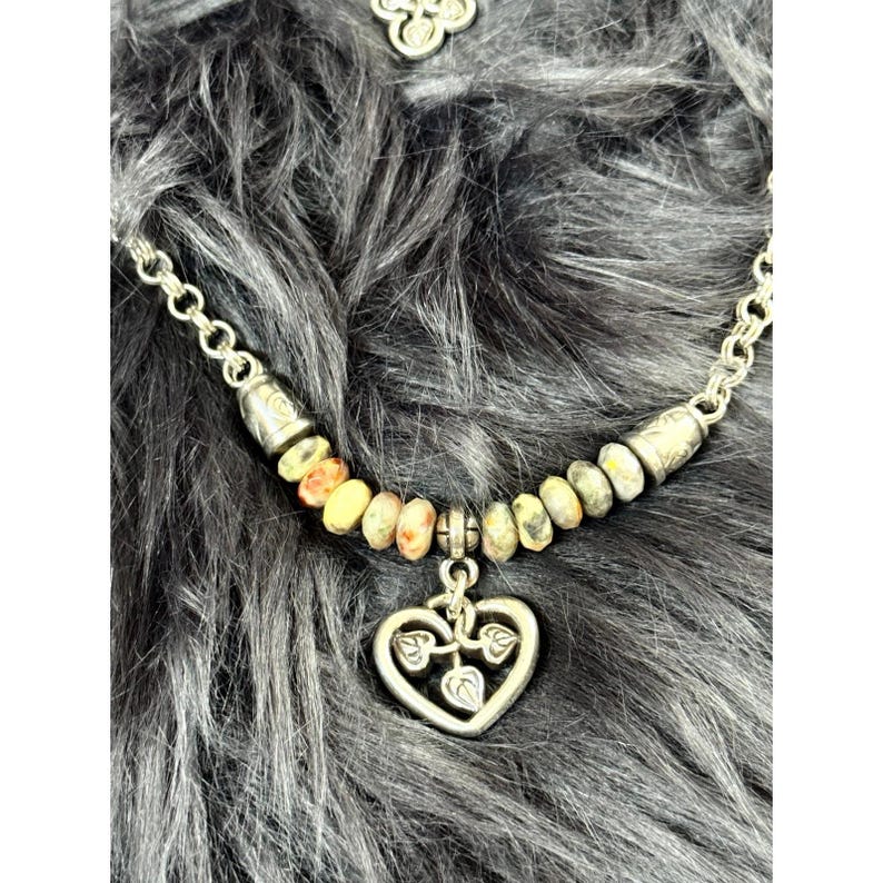 May include: A silver-toned necklace with a heart pendant featuring leaf designs. The necklace has a chain with small, round links and a beaded section with yellow, orange, and gray stones. The background is a fuzzy, gray surface.