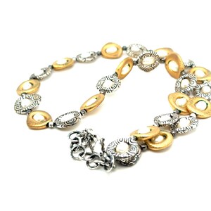 Puede incluir: Un collar plateado y dorado con una variedad de formas, incluyendo dise&ntilde;os ovalados, redondos y circulares. El collar presenta una mezcla de texturas y acabados, con un cierre de broche. La joya se exhibe sobre un fondo blanco.