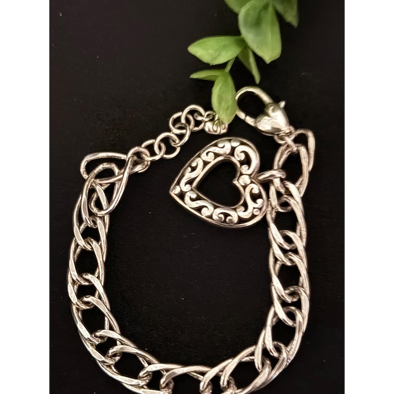 Op de afbeelding: Zilverkleurige armband met een hartvormige bedel met een open ontwerp. De bedel is verbonden met een schakelarmband. De armband wordt getoond op een donkere ondergrond, met een takje groene bladeren op de achtergrond.