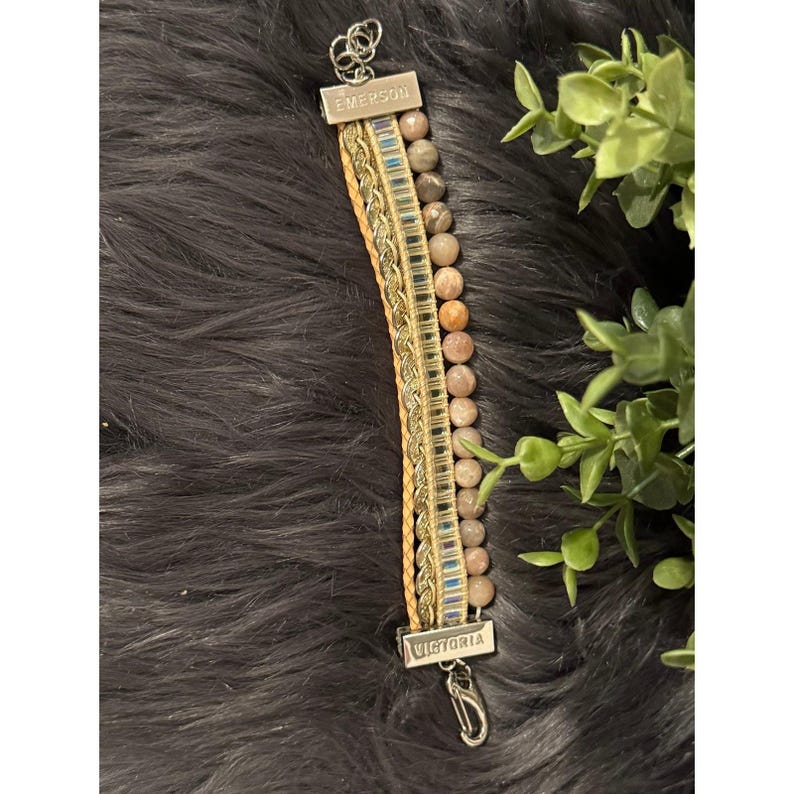 Op de afbeelding: Een armband met meerdere strengen met een mix van materialen en texturen. De armband bevat een streng ronde, lichtbruine kralen, geweven leer en een goudkleurige ketting. De armband heeft metalen staven met de tekst "Emerson" en "Victoria".