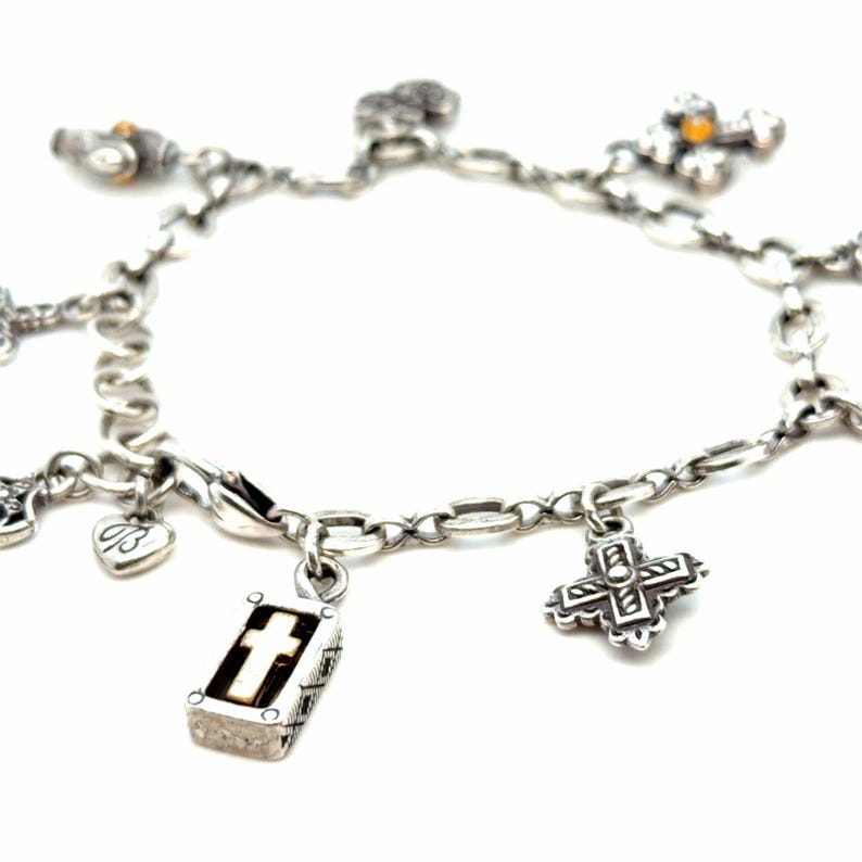May include: A silver charm bracelet with various religious-themed charms. Charms include crosses, a heart with the initials "B", and a small book with a cross. The bracelet is on a white background.
