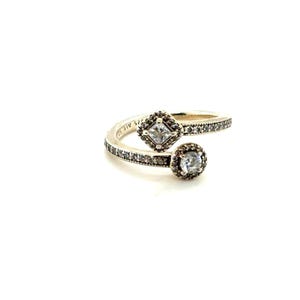 May include: A silver bypass ring featuring a square-cut clear stone in a halo setting on one end and a round-cut clear stone in a halo setting on the other. The band is encrusted with small clear stones and is engraved with the text S925 ALE 52.