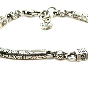 Puede incluir: Primer plano de una pulsera de plata con intrincados dise&ntilde;os grabados. La pulsera presenta un dije en forma de coraz&oacute;n y varias cuentas y eslabones cil&iacute;ndricos decorativos. El cierre es visible, sobre un fondo blanco.