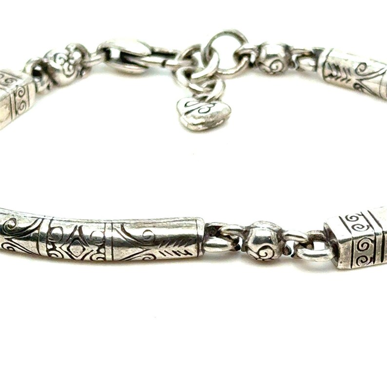 Puede incluir: Primer plano de una pulsera de plata con intrincados dise&ntilde;os grabados. La pulsera presenta un dije en forma de coraz&oacute;n y varias cuentas y eslabones cil&iacute;ndricos decorativos. El cierre es visible, sobre un fondo blanco.