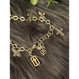 May include: Silver-toned charm bracelet with various religious-themed charms. Charms include crosses, a four-leaf clover, and a shield. The bracelet is displayed on a dark, textured surface.