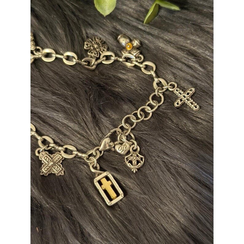 May include: Silver-toned charm bracelet with various religious-themed charms. Charms include crosses, a four-leaf clover, and a shield. The bracelet is displayed on a dark, textured surface.
