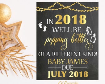 May include: Gold glitter and star decorations on a white background with a black chalkboard sign announcing a baby is due in July 2018. The sign reads "In 2018 we'll be popping bottles of a different kind! Baby James due July 2018".