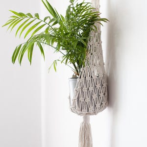 Op de afbeelding: Een crème macramé plantenhouder met een groene plant. De geweven houder hangt aan een witte muur, met een kwast aan de onderkant. De plant heeft lange, dunne groene bladeren, wat een natuurlijke uitstraling creëert.