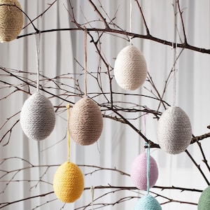 Può includere: Una collezione di sei uova di Pasqua in lana fatte a mano appese a un ramo. Le uova sono di vari colori, tra cui beige, marrone chiaro, giallo, rosa e grigio.