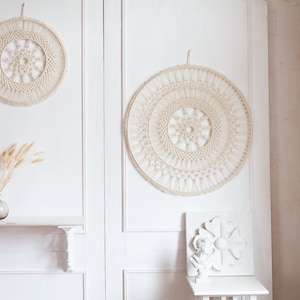 May include: Two white macrame wall hangings with a circular design, one larger than the other, hanging on a white wall. A vase with dried grass is on a white shelf below the smaller wall hanging.