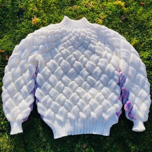 May include: White knitted sweater with a textured diamond pattern and pink and purple accents on the sleeves.