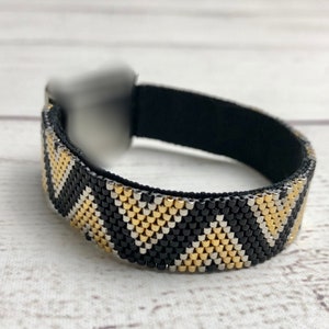 Op de afbeelding: Een zwarte, gouden en zilveren kralen armband met een geometrisch patroon. De armband is gemaakt met kleine, vierkante kralen en heeft een zwart elastiek.