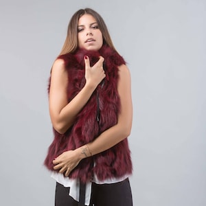 May include: A sleeveless, burgundy faux fur vest with a black zipper closure. The vest has a plush texture and a high collar. The model is wearing a white top underneath. The background is a solid, light gray.