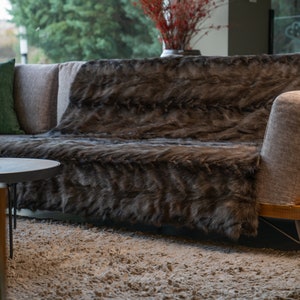 Op de afbeelding: Een bruine nep bont deken gedrapeerd over een moderne bank met beige kussens. De bank staat in een woonkamer met een kleine ronde tafel en een tapijt.