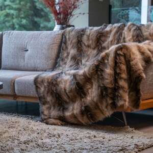 Op de afbeelding: Een grijze bank met een houten basis en een faux fur plaid. De deken heeft een mix van bruine en grijze tinten, waardoor een textuur ontstaat. Een lichtgekleurd tapijt staat op de voorgrond.