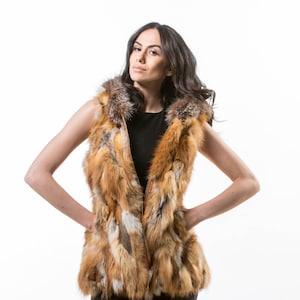 May include: A sleeveless fur vest in shades of brown, orange, and cream. The vest has a zip-up front and a collar with a mix of brown and grey fur. The model is wearing a black top underneath. The vest is a statement piece.