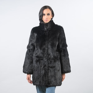 May include: A black fur coat with a high collar and long sleeves. The coat has a mid-thigh length and a slightly flared silhouette. The model is wearing blue jeans. The coat appears to be made of a soft, plush material.
