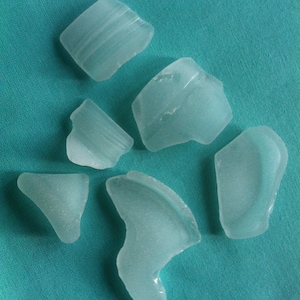 Puede incluir: Colección de seis piezas de vidrio de mar de diversas formas y tamaños. El vidrio de mar es de un color azul-verdoso pálido y translúcido, con sutiles estrías en algunas piezas. Están dispuestas sobre una tela de color turquesa.