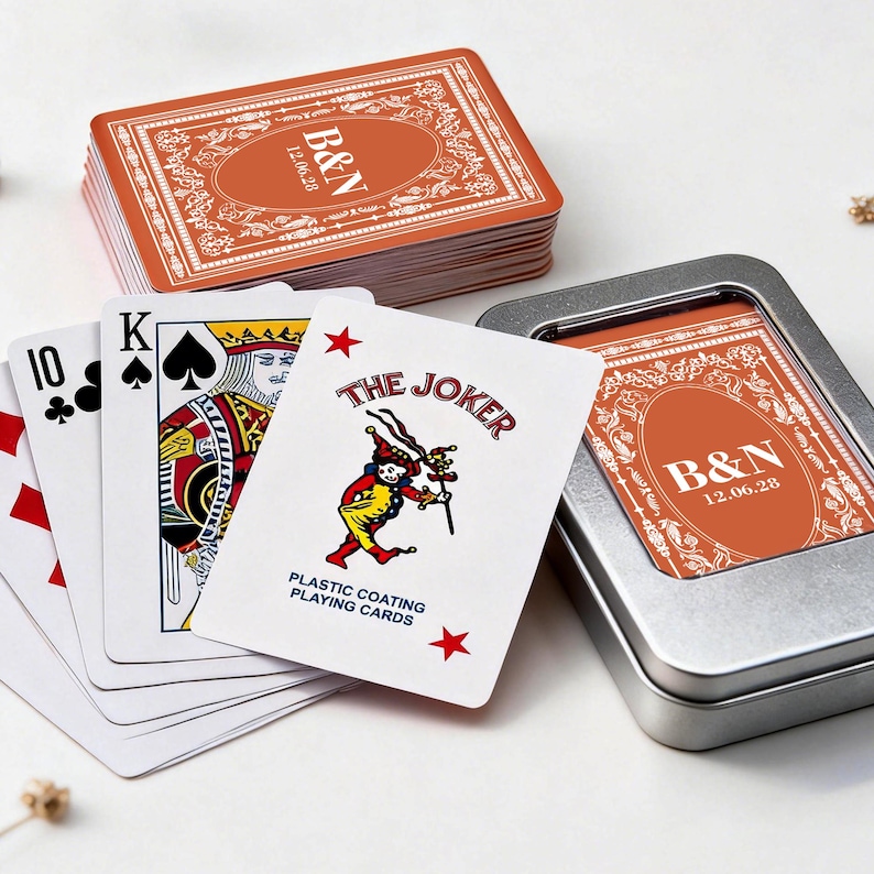 May include: A deck of playing cards with a personalized orange and white design, featuring the initials "B&N" and the date "12.06.28". The cards are displayed with a metal tin and a few cards fanned out, including the Joker card.