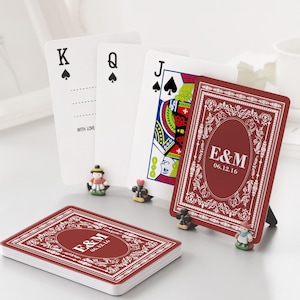 May include: A deck of playing cards with a red and white decorative design. The cards are personalized with the initials "E&M" and the date "06.12.16". The image also shows the King, Queen, and Jack of Spades.