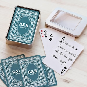 May include: A deck of teal playing cards in a silver tin. The cards feature white ornate designs and the initials "B&R" with the date "12.02.22". Several cards are displayed, including an Ace of Spades with handwritten text.