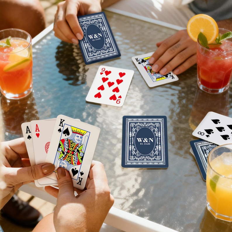 May include: A game of cards with personalized navy blue playing cards featuring the initials "W&N" and the date "01.15.26". The cards are on a glass table with cocktails.
