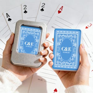 May include: A deck of playing cards with a blue and white design, held in a silver tin. The cards feature the initials "C&E" and the date "12.03.22". Several playing cards are scattered around, including the Ace of Spades and the Ace of Hearts.