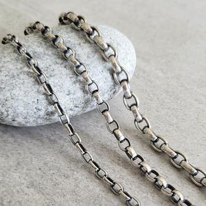 May include: Three silver chains of different thicknesses, laid out on a gray stone background.