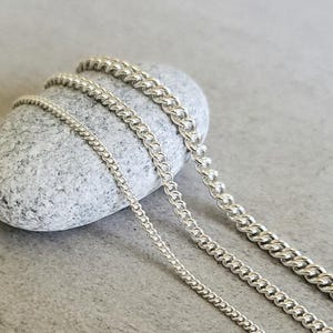 May include: Four silver chains of different thicknesses, arranged on a gray stone background.