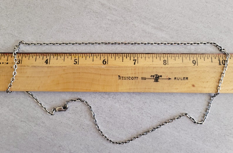 May include: A silver chain necklace measuring 9 inches long, laid out on a wooden ruler with the Westcott brand name and the word "ruler" printed on it.