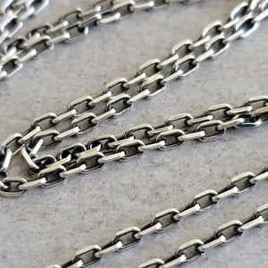 May include: Close-up of a silver chain necklace with a box chain design. The chain is made of small, rectangular links that are connected together.