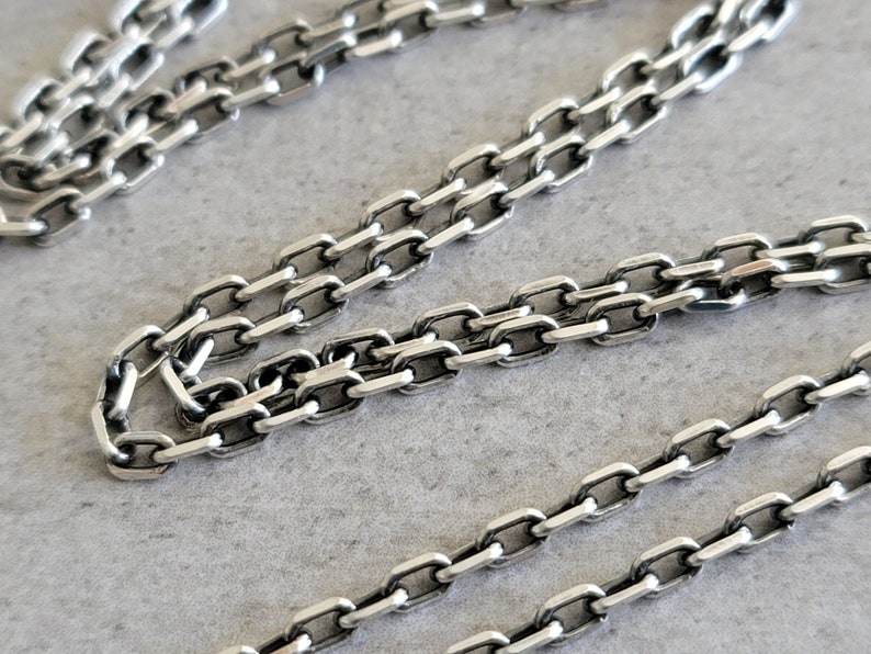 May include: Close-up of a silver chain necklace with a box chain design. The chain is made of small, rectangular links that are connected together.