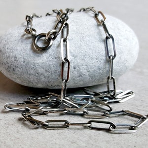 May include: A silver chain necklace with a simple, rectangular link design. The chain is lying on a gray stone.