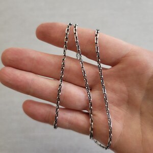 May include: A silver chain necklace with a box chain design. The chain is laying on a hand.