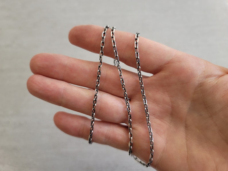 May include: A silver chain necklace with a box chain design. The chain is laying on a hand.