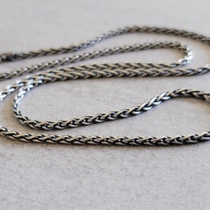 May include: A silver-colored, braided chain necklace. The necklace is laid out on a light gray surface. The chain has a textured, woven appearance, with a clasp at one end. The overall design is simple and elegant.