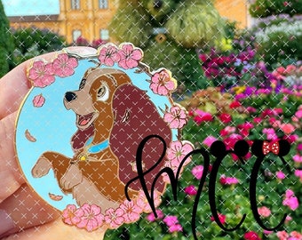 May include: A round enamel pin featuring a cartoon dog with brown fur, a blue collar, and a gold tag. The dog is surrounded by pink cherry blossoms on a light blue background. The pin has a gold border.