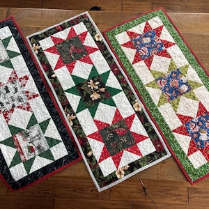 May include: Three rectangular quilted table runners with star patterns. Each runner features a central star design in red, green, and white, with varying patterns and border colors. The runners are displayed on a wooden surface.