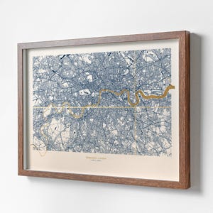 May include: A framed map of Greenwich, London, showing a detailed blue and white street plan with a gold-coloured outline of the River Thames. The print is set in a brown wooden frame.