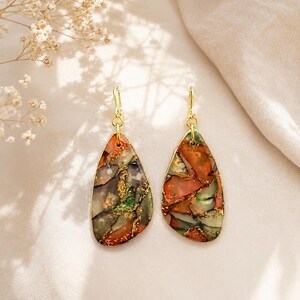 May include: A pair of teardrop-shaped dangle earrings featuring a multi-colored marbled pattern in shades of deep red, forest green, and grey, accented with flecks of gold leaf. The earrings are suspended from simple gold-toned metal hooks.