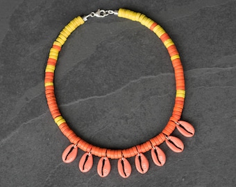 May include: A necklace featuring a strand of small, flat, circular beads in alternating yellow and orange colours. The lower section of the necklace is adorned with seven dangling, oval-shaped, coral-coloured cowrie shells attached with silver-toned metal rings.