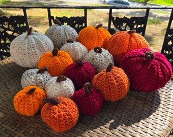 May include: A collection of crocheted pumpkins in various sizes and colors, including white, orange, and red. The pumpkins are arranged on a woven wicker table.