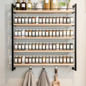 May include: A black metal spice rack with five wooden shelves, filled with glass jars of spices. The rack is mounted on a white wall with two towels hanging on hooks below it.