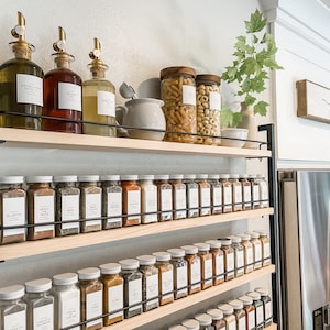 May include: A black metal shelf unit with four wooden shelves holds a collection of glass spice jars and bottles. The shelves are filled with various spices, including garlic, onion, and pepper. The bottles are filled with olive oil and vinegar. The shelf unit is mounted on a white wall. The word "gather" is written on a wooden sign above the shelf unit.