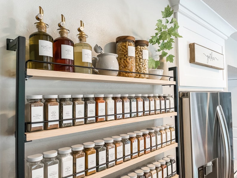 May include: A black metal shelf unit with four wooden shelves holds a collection of glass spice jars and bottles. The shelves are filled with various spices, including garlic, onion, and pepper. The bottles are filled with olive oil and vinegar. The shelf unit is mounted on a white wall. The word "gather" is written on a wooden sign above the shelf unit.