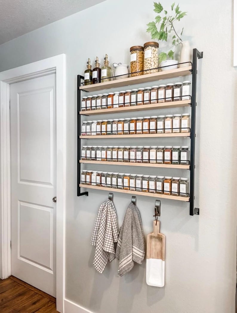 May include: A wall-mounted spice rack with five shelves, filled with glass jars of spices. The rack is made of black metal and wood. There are two towels hanging on hooks below the rack, and a wooden cutting board is hanging on a hook to the right.