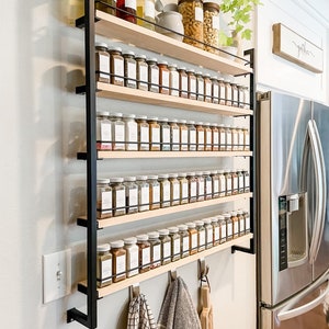 May include: A black metal spice rack with five wooden shelves, filled with glass jars of spices. The rack is mounted on a white wall and has hooks for hanging towels. The text "gather" is visible on a wooden sign above the refrigerator.