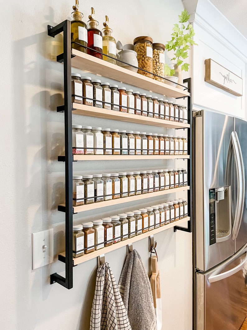 May include: A black metal spice rack with five wooden shelves, filled with glass jars of spices. The rack is mounted on a white wall and has hooks for hanging towels. The text "gather" is visible on a wooden sign above the refrigerator.