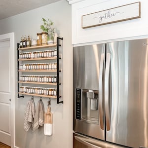 May include: A stainless steel refrigerator with a water dispenser and a wooden sign above it that reads "gather". A spice rack with many glass jars of spices is mounted on the wall to the left of the refrigerator.